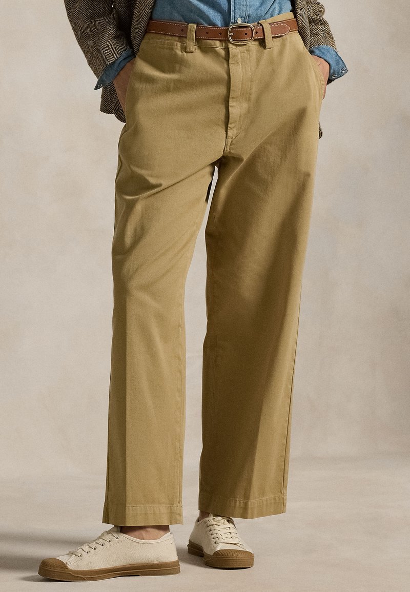 Personne portant un pantalon beige droit, une ceinture marron, une chemise en denim, un blazer à chevrons et des baskets blanches en toile, les mains dans les poches.
