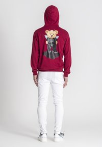 Bordo dukserica s grafikom medvjeda s krilima i crnim outfitom na leđima. Uparena s bijelim uskim trapericama i neutralnim obućom.