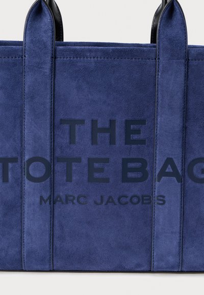 Bolso tote de ante azul marino con texto audaz "THE TOTE BAG" y "MARC JACOBS" en un color oscuro contrastante. Diseño sencillo con asas resistentes.