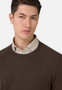 Jeune homme aux cheveux courts et foncés portant une chemise beige à col sous un pull marron à col rond, fond neutre.