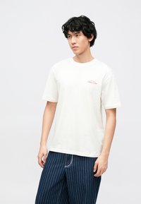 Hvid bomulds t-shirt med korte ærmer, med et lille rødt broderet logo på venstre bryst. Parret med blå bukser med tynde striber.