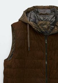 Veste sans manches matelassée marron avec capuche, fermeture éclair devant et cordon de serrage, montrant une doublure intérieure brillante et une étiquette de marque à l'intérieur du col.