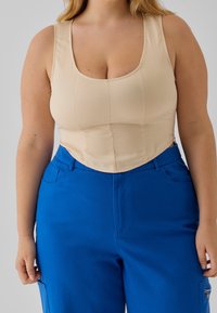 Béžový přiléhavý top s vykrojeným výstřihem a strukturovanými švy; doplněný modrými kalhotami se širokými nohavicemi, bočními kapsami a předním zipem.