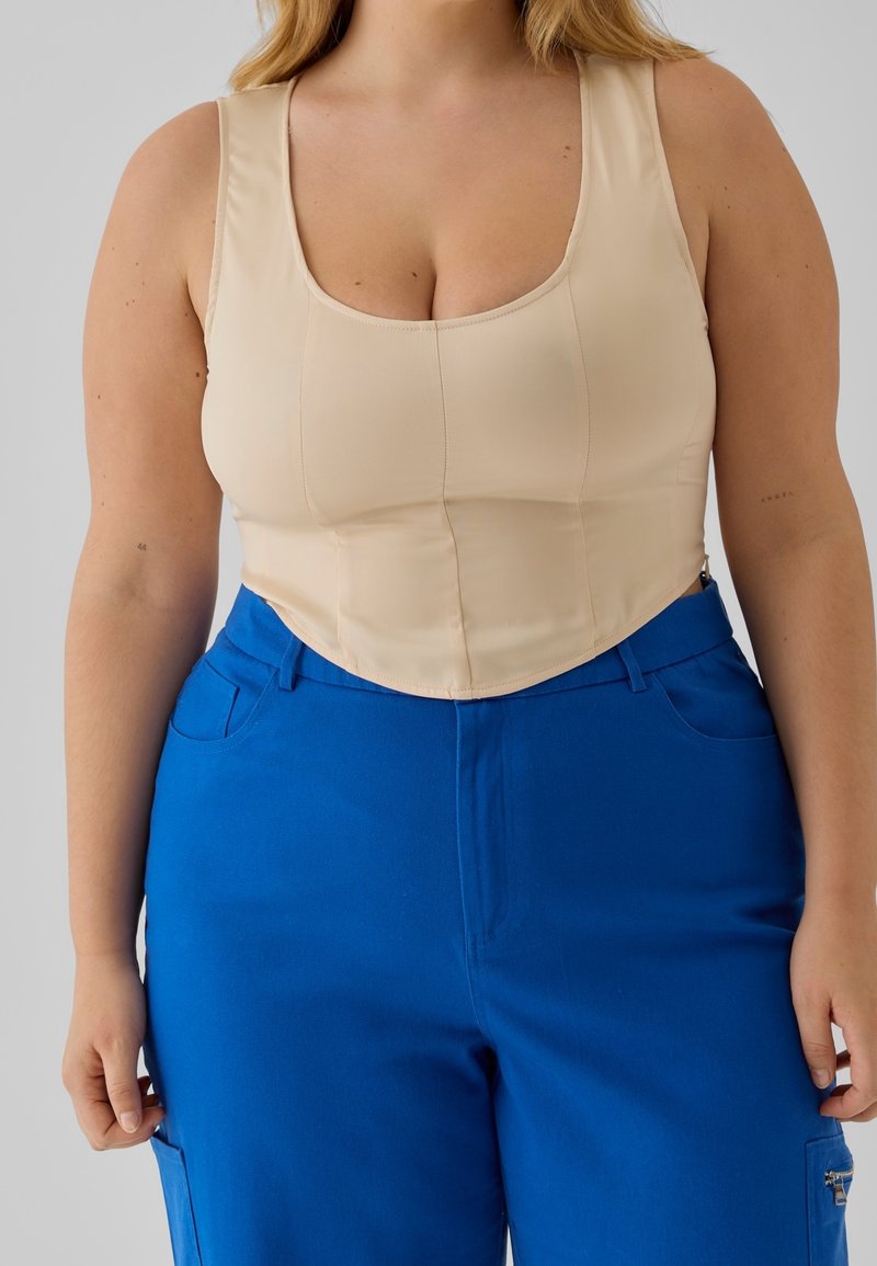 Béžový přiléhavý top s vykrojeným výstřihem a strukturovanými švy; doplněný modrými kalhotami se širokými nohavicemi, bočními kapsami a předním zipem.