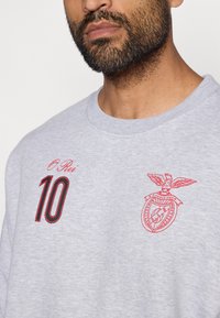 Sudadera gris con detalles bordados en rojo, que presenta el número "10" y un escudo con un águila. Cuello redondo y textura suave.