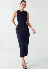 Navyblaues ärmelloses Kleid mit einem Cowl-Ausschnitt, einem gebundenen Taillendetail und Midi-Länge, gefertigt aus glattem Stoff. Getragen mit flachen schwarzen Sandalen.