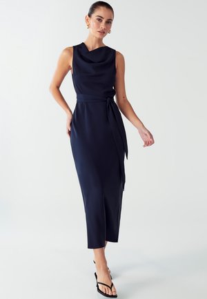 Navy blue sleeveless dress with a cowl neckline, a tied waist detail, and a midi length, made from smooth fabric. Worn with flat black sandals.