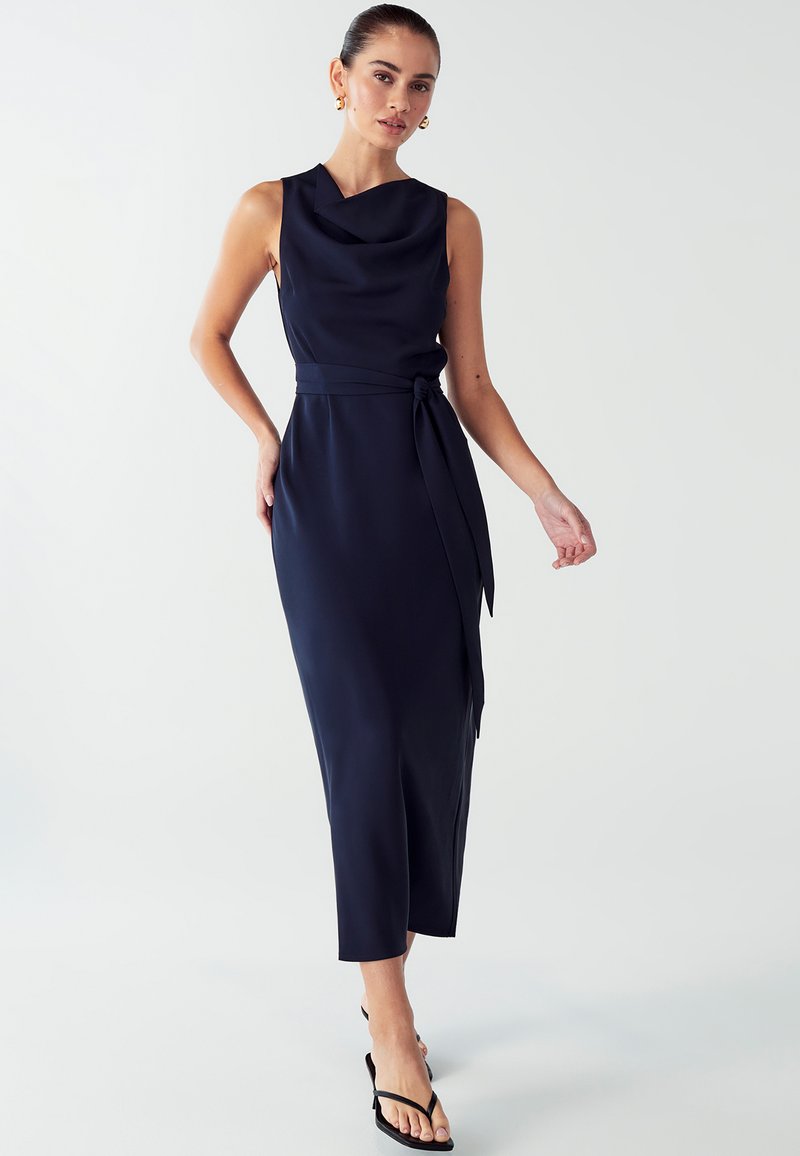 Navyblaues ärmelloses Kleid mit einem Cowl-Ausschnitt, einem gebundenen Taillendetail und Midi-Länge, gefertigt aus glattem Stoff. Getragen mit flachen schwarzen Sandalen.