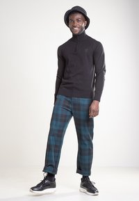 Czarny sweter z dzianiny z długim rękawem i półzamkiem; spodnie w kratę w odcieniach niebieskiego i zielonego; czarne trampki; czarny, okrągły kapelusz; postawa stojąca na jasnym tle.