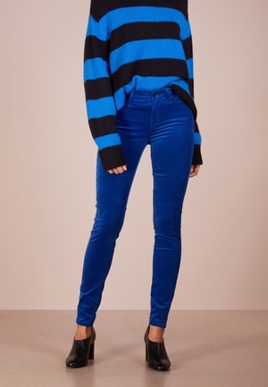 Persona que lleva pantalones pitillo de terciopelo azul brillante, botines negros de tacón alto y un suéter a rayas azul y negro.