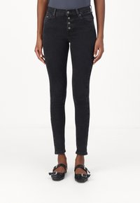 Černé vysoký pas úzké džíny s pěti knoflíkovým zapínáním, předními kapsami a úzkým střihem. Kombinovány s černými botami se čtvercovou špičkou a kotníkovými pásky.