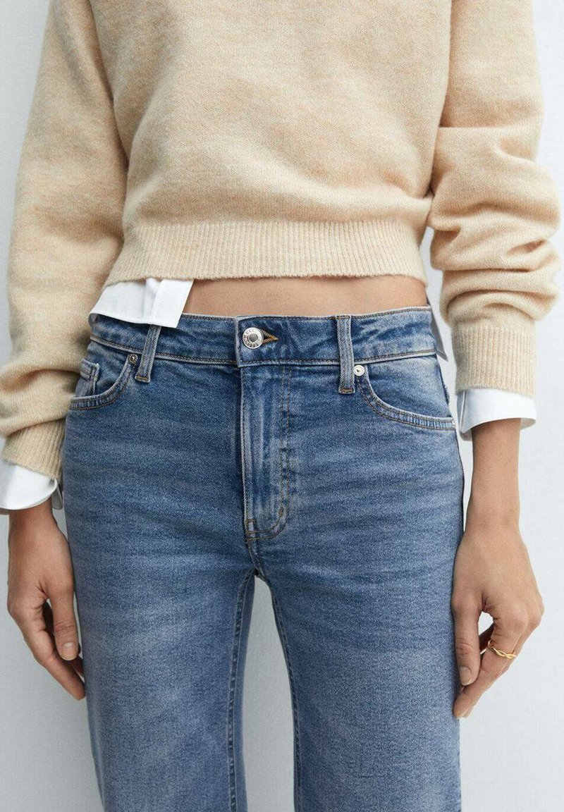 Licht beige gebreid sweatervest over een wit shirt, gecombineerd met hoogvrouwelijke blauwe denim jeans met subtiele vervaging en een messing knoop.