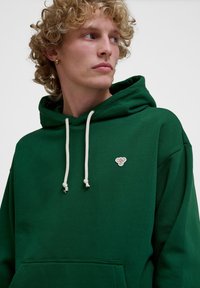 Zielona bawełniana bluza z kapturem z przednią kieszenią, białymi sznurkami do ściągania oraz małym logo na piersiach. Gładka tekstura, luźny krój.