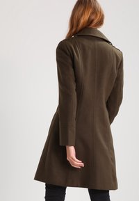 Abrigo de lana ajustado en verde oliva, con cuello, mangas largas y dobladillo acampanado. Presenta detalles en los hombros y costuras verticales.
