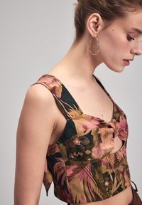 Blomster mønstret cropped top med brede skulderstropper, der har et udskåret design. Farverne inkluderer dybe grønne nuancer, rosa og brune.