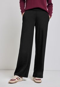 Zwarte wide-leg broek van zachte stof, met zijzakken en een relaxte pasvorm, gecombineerd met beige en bordeauxrode sneakers.