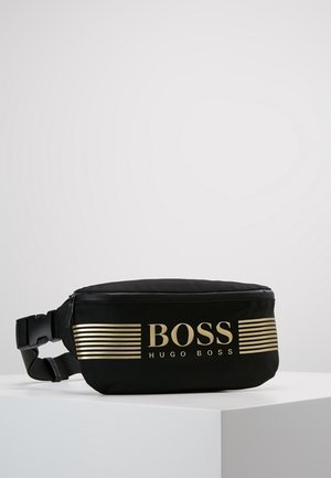 Musta Hugo Bossin vyölaukku, jossa on kultaiset vaaka raidat ja logo, säädettävä olkahihna sekä vetoketjullinen pääosasto valkoisella pinnalla.