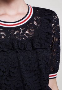 Schwarzes Spitzentop mit einem rüschierten Vorderteil, rundem Halsausschnitt mit rot-weiß gestreiftem Saum und kurzen Puffärmeln.