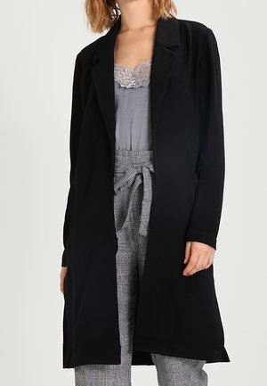 Cappotto lungo nero con un design frontale aperto, taglio dritto e stile minimalista. Presenta un tessuto morbido, abbinato a un top in pizzo grigio e pantaloni a fantasia.