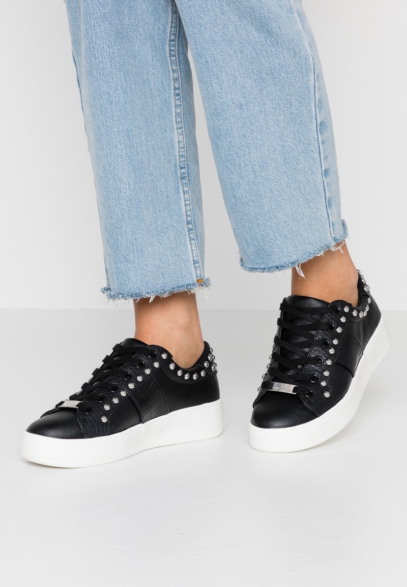 Musta keinonahkakengät, joissa on hopeiset niitit kauluksessa, teksturoidut sivupaneelit ja paksu valkoinen pohja. Yhdistetty vaaleansinisiin lyhyisiin farkkuihin.