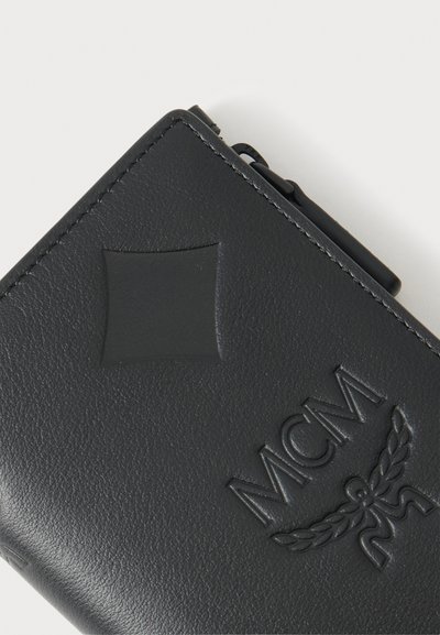 Portefeuille en cuir noir avec logo MCM embossé et un détail carré subtil près du coin supérieur, bords cousus visibles.