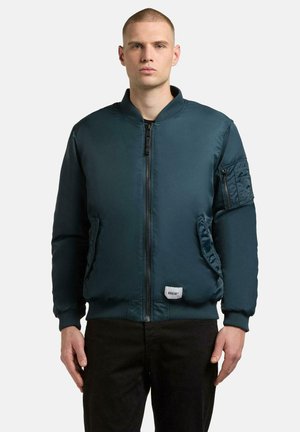 Giacca bomber di un verde scuro con zip frontale, due tasche laterali e polsini elasticizzati. Presenta una tasca a toppa sulla manica.