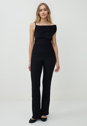 Vrouw staat in een textuur zwarte top met één schouderband en bijpassende flared broek tegen een eenvoudige lichte achtergrond.