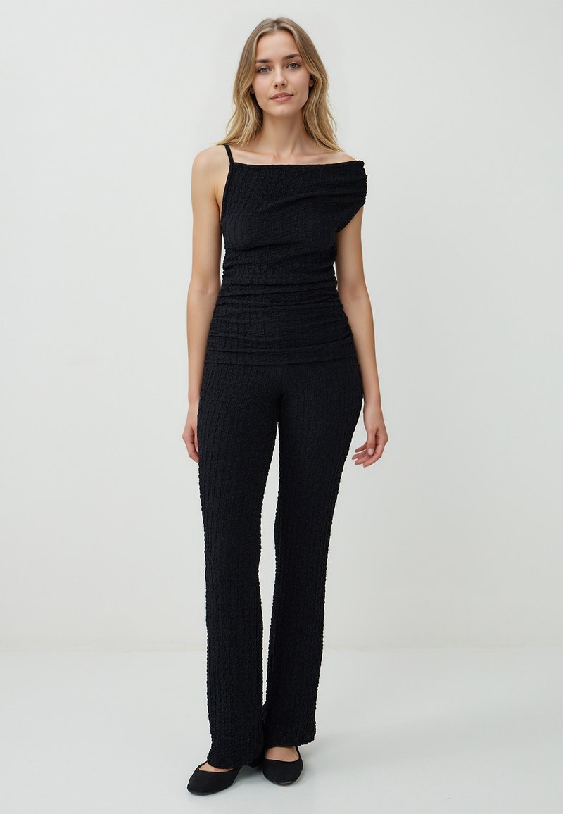 Femme debout dans un haut noir texturé à une épaule avec une sangle et un pantalon évasé assorti, sur un fond clair uni.