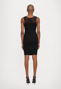 Black fitted dress with a sheer mesh top panel, knee-length. Worn with black high-heeled sandals, showing a streamlined silhouette.