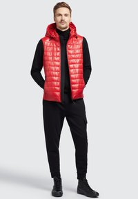 Rød, quiltet puffervest med hætte, lynlås lukning, båret over en sort turtleneck, parret med sorte joggers og højhælede sneakers.