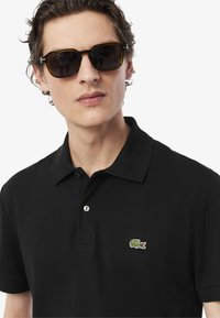 Camiseta polo negra con una tapeta de dos botones y un pequeño logo de cocodrilo verde. El modelo lleva gafas de sol oscuras con un marco cuadrado.