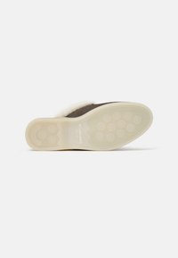 Zapatilla de ante marrón con forro de piel sintética crema. La suela de goma blanca presenta patrones de agarre circulares, un logotipo en relieve y bordes texturizados.
