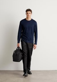Navy long-sleeve shirt, black textured pants, and black leather shoes. Black bag with handles. Model walking on a light gray floor.