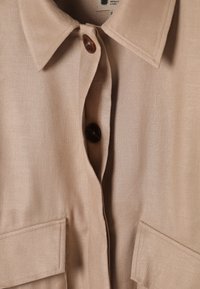 Gros plan du col d'une chemise beige avec deux boutons marron et deux poches avant, montrant la texture du tissu et les détails des coutures.