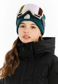 Zwarte pufferjack met hoge kraag, voorzien van een turquoise brei-beanie en spiegeltinten skibril die erop rust. Zachte, gladde textuur.