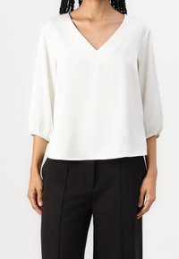Bílá, lehká blůza s výstřihem do V, tříčtvrtečním nadýchaným rukávem a elastickými manžetami. Rovný lem, hladká textura, spárovaná s černými kalhotami.