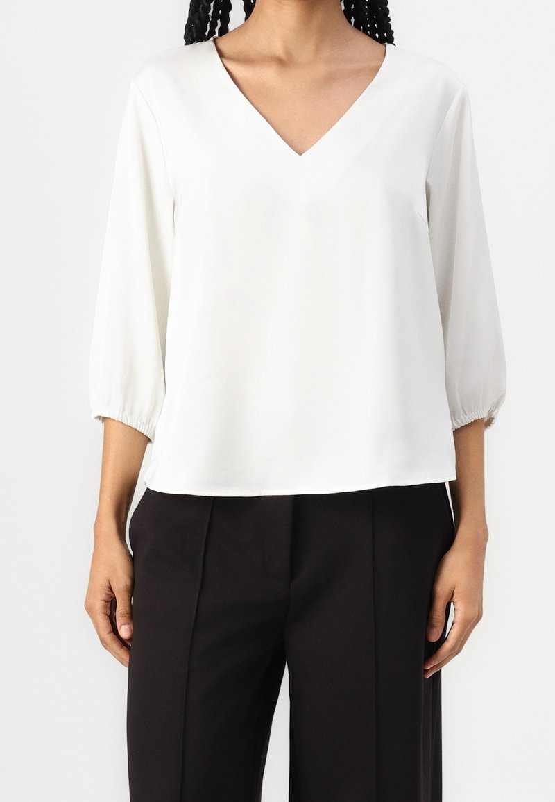 Bílá, lehká blůza s výstřihem do V, tříčtvrtečním nadýchaným rukávem a elastickými manžetami. Rovný lem, hladká textura, spárovaná s černými kalhotami.