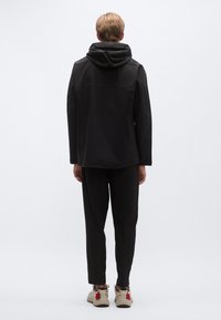 Fekete, kapucnis dzseki laza szabással, fekete nadrággal és bézs sportcipővel, rajta piros részletek. Hátsó nézet, sima textúrával és tiszta vonalakkal.