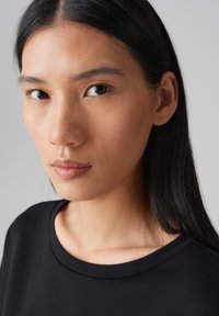 Schwarzes, T-Shirt mit Rundhalsausschnitt und glatter Textur, umrahmt von langen, glatten, schwarzen Haaren. Natürlicher Look mit minimalem Make-up und klarer Haut.