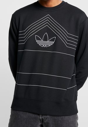 Sweat-shirt noir avec des lignes géométriques blanches et le logo trefoil d'Adidas brodé sur la poitrine, porté par une personne avec les mains dans le dos.