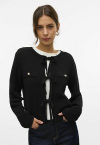 Cardigan en tricot noir avec un col rond, trois nœuds à l'avant et deux poches poitrine boutonnées. La texture est douce et polyvalente.