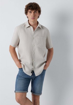 Junger Mann mit lockigem Haar, der ein beiges kurzärmeliges Hemd mit Knöpfen und hochgekrempelte Jeansshorts trägt, die Hände in den Taschen.