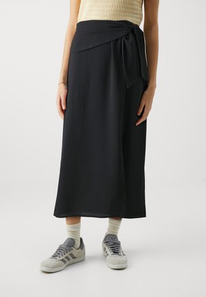 Svart omslagskjørt laget av glatt stoff, med en knytedetalj foran og en lang, flytende form. Kombinert med grå sneakers og beige sokker.