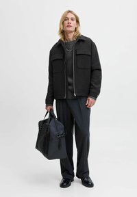 Sort zip-up jakke med to frontlommer, parret med en mørk sweater og grå plisserede bukser. Modellen holder en sort lædertaske.