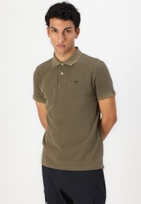 Olivgrünes Poloshirt aus weichem Material, mit zwei Knöpfen am Kragen und hochgeschlagenen Ärmeln. Verfügt über ein dezentes Logo auf der Brust.
