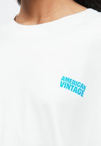 Κοντινό πλάνο ενός ατόμου που φοράει ένα λευκό πουκάμισο με μπλε κείμενο που αναγράφει "AMERICAN VINTAGE" στο αριστερό στήθος.