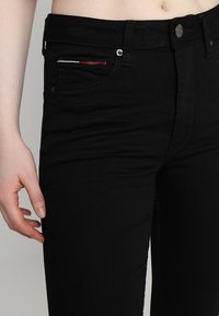 Vaqueros de mezclilla negros con un corte ajustado, con cierre de botón, bolsillo delantero con costura decorativa en rojo y blanco, y una textura sutil.