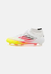 Des chaussures de football blanches avec un design de cheville haute, présentant des accents noirs, une semelle dégradée allant du jaune au rose, et un dessus texturé pour une bonne adhérence.