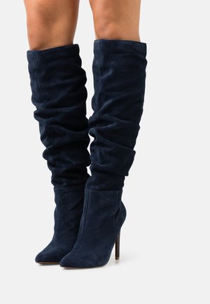 Bottes en daim bleu marine montant au-dessus du genou avec un design décontracté et un bout pointu, dotées d'un talon aiguïlle haut et d'une texture douce.
