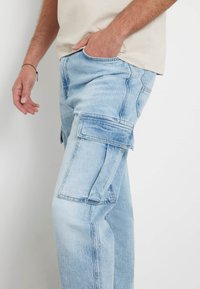 Pantaloni cargo in denim azzurro chiaro con numerose tasche laterali, vestibilità rilassata e aree sbiadite. Il tessuto è testurizzato e presenta dettagli di cucitura tradizionali.